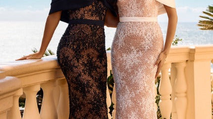 Two people wearing formal lace evening gowns pose together on a seaside terrace beside a stone balustrade in daylight